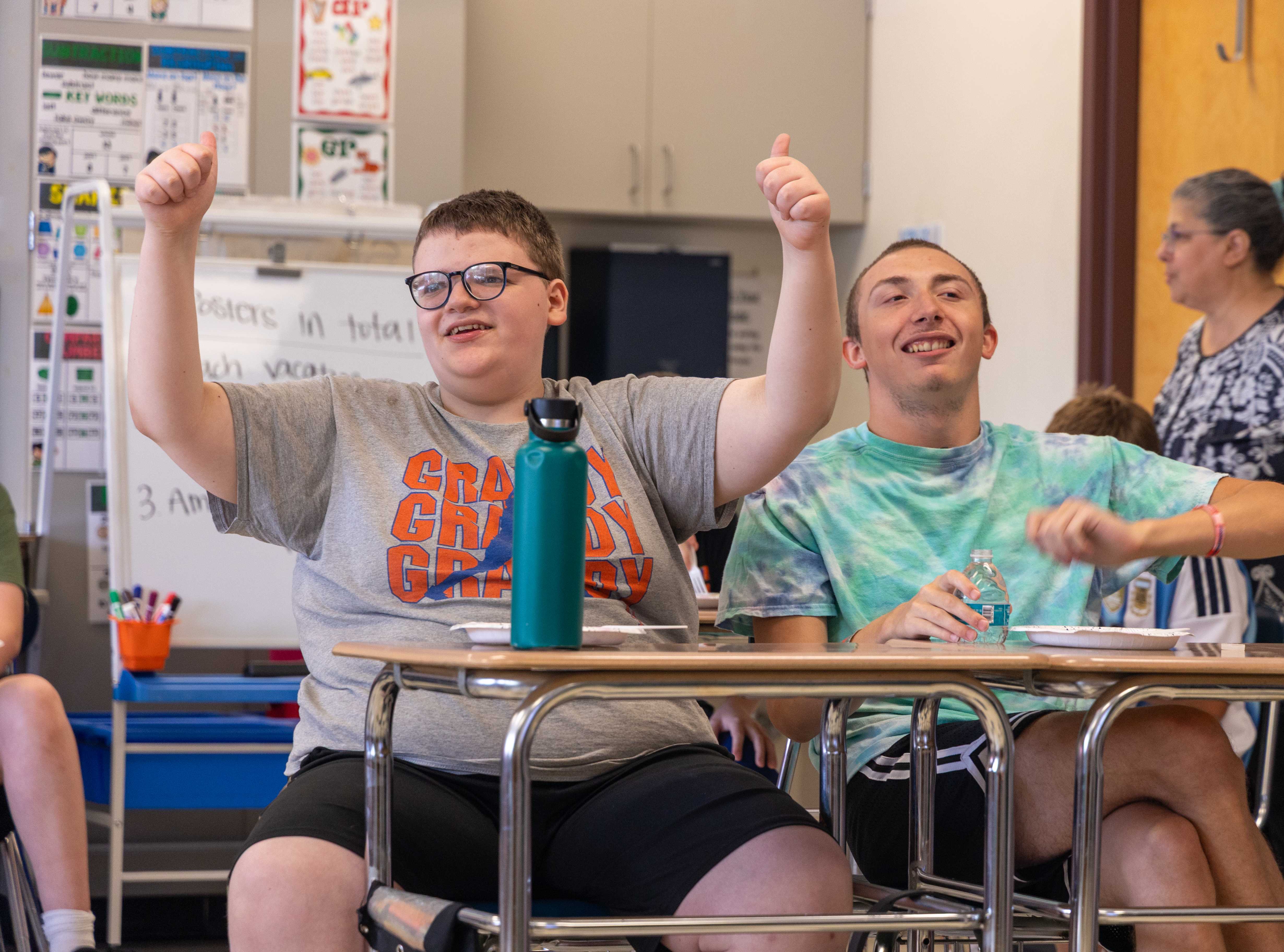 two students smile at their desks, one holding two thumbs up